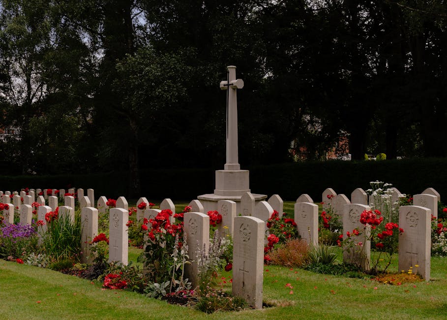 A peaceful cemetery scene featuring a row of white gravestones, each engraved with a floral motif or cross, arranged in neat lines on well-maintained green grass. In the center background stands a whi