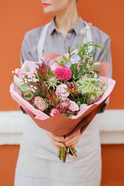 A person wearing a light grey shirt and white apron holds a large bouquet of fresh flowers, wrapped in pink and brown paper, in front of an orange wall. The bouquet features pink and white carnations,
