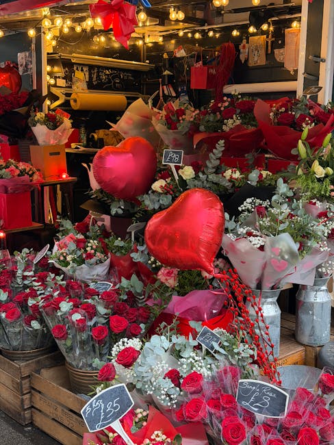 A vibrant display of floral arrangements at a flower shop, featuring predominantly red and white roses, some wrapped in pink tissue paper and others in transparent wrapping. The flowers are arranged i