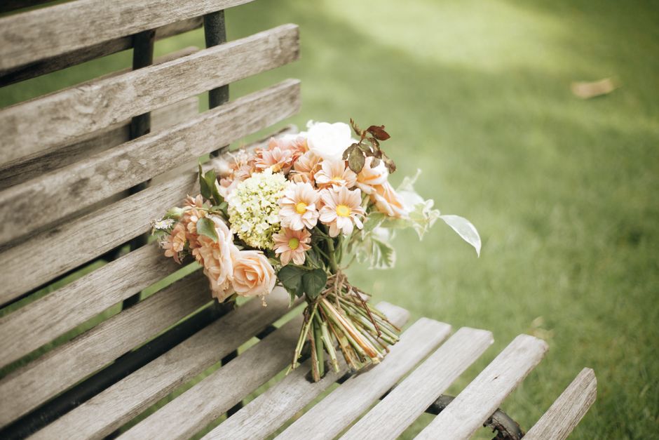 A bouquet of fresh flowers resting on a weathered wooden park bench, featuring soft peach roses, light pink daisies with yellow centers, white and green hydrangea clusters, and assorted greenery with 